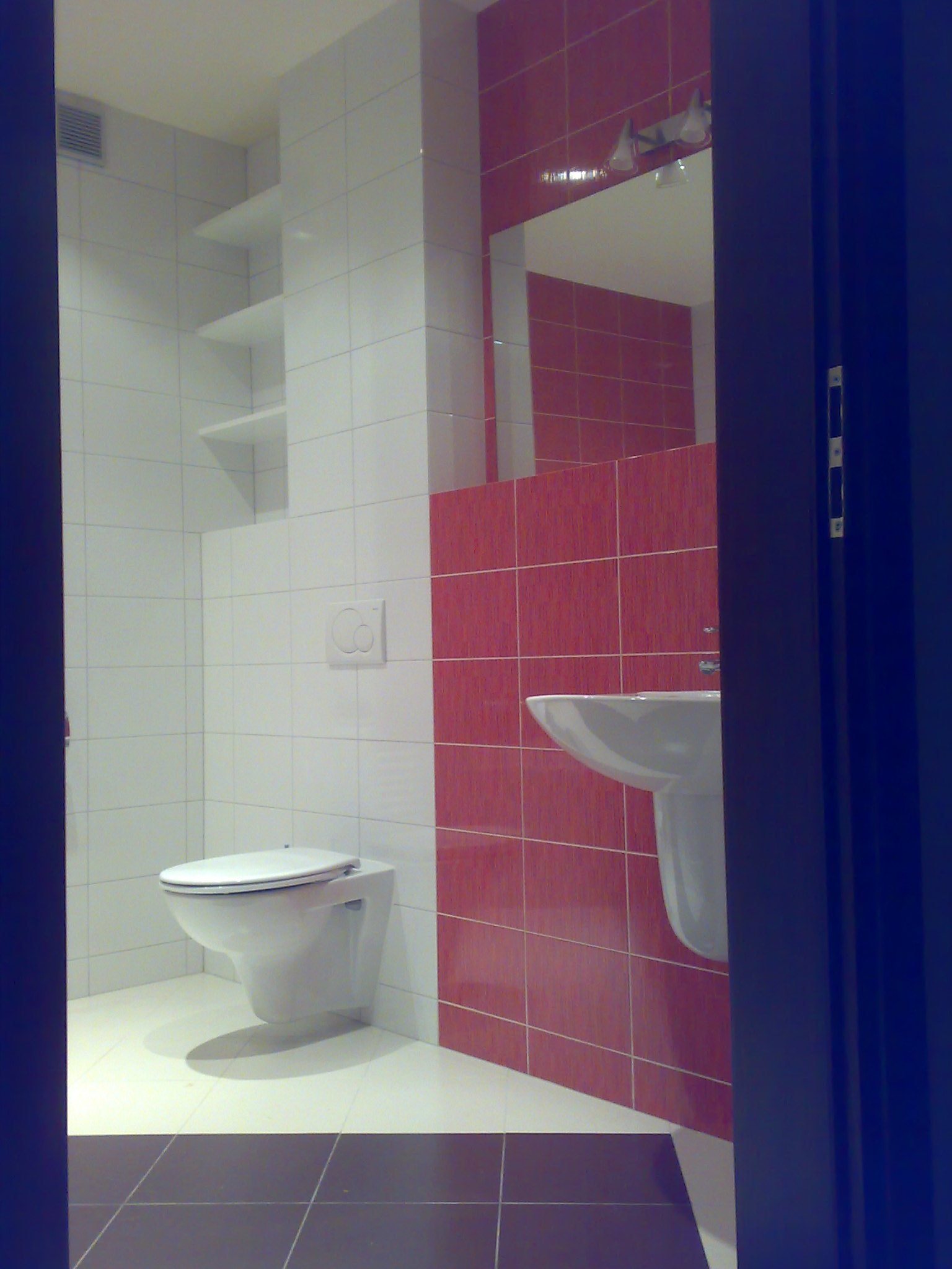 Wykończona łazienka z biało-czerwonymi płytkami, wiszącą toaletą, umywalką na postumencie i półkami, widok zza uchylonych drzwi.